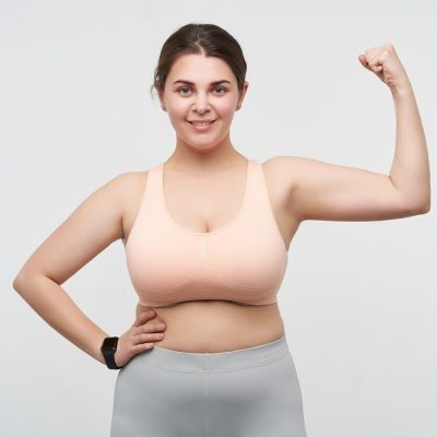 Indoor photo of young cheerful brunette chubby female in sporty clothes keeping hand raised while demonstrating her power, isolated over white background. Concept of weight loss