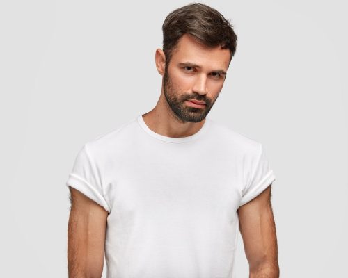 Serious muscular young male with dark stubble, hair, dressed in casual white t shirt, has muscular body, listens attentively something, isolated over white background. Unshaven guy stands indoor